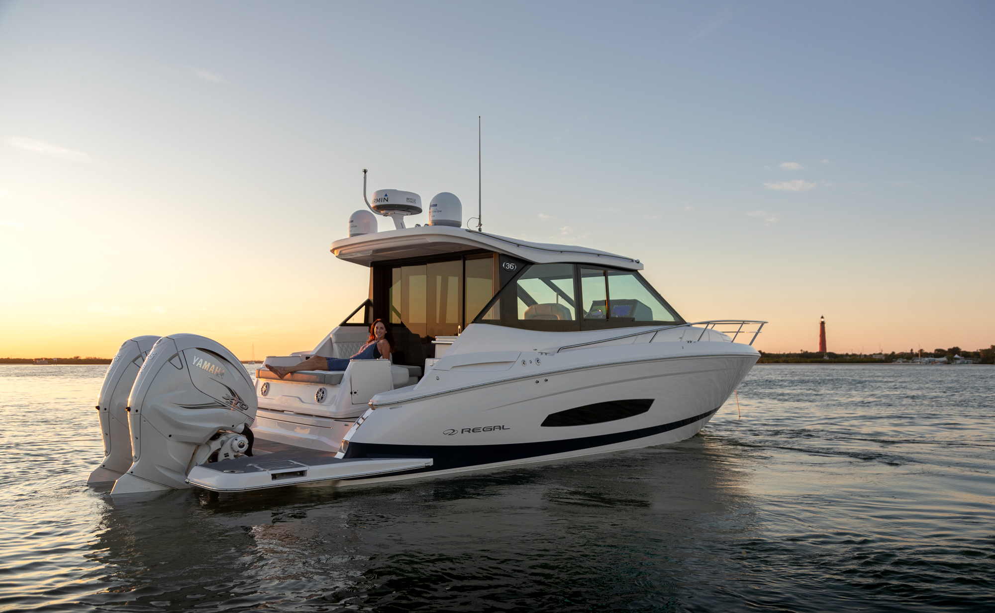A Regal 36 coupe at rest on calm water at golden-hour sunset, with a guest lounging on the aft sunpad and a red lighthouse on the distant shoreline.